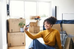 Woman With Mobile Phone Moving Into New Home Making Video Call Surrounded By Boxes